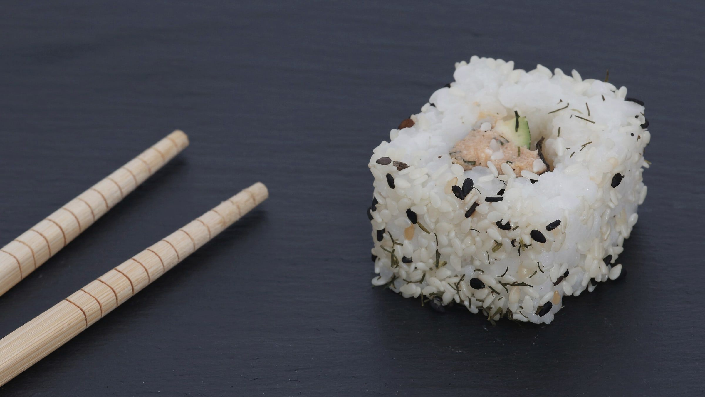 Un uramaki solo sobre pizarra oscura, junto a un par de palillos de madera